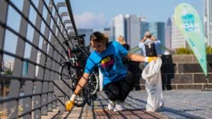 Eine junge Frau in blauem World-Cleanup-Day-T-Shirt hebt am Mainufer Abfall vom Boden auf, im Hintergrund die Frankfurter Skyline und eine Beachflag mit der Aufschrift Mach mit.