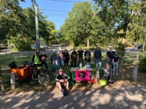 Eine Gruppe Erwachsener steht und sitzt an einem Tisch mit World-Cleanup-Day-Plakat an einer baumbestandenen Straße, daneben ein Lastenrad.