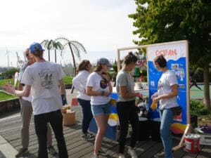 Mehrere Helferinnen in World-Cleanup-Day-Shirts stehen an einem Stand mit einer Ocean-Stellwand, im Hintergrund Palmen und Hafenkräne.