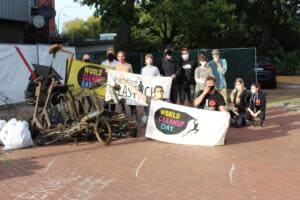 Eine Gruppe mit Masken steht und kniet hinter Bannern von World Cleanup Day und Initiative Plastich, davor Sperrmüll, Einkaufswagen und ein altes Fahrrad.