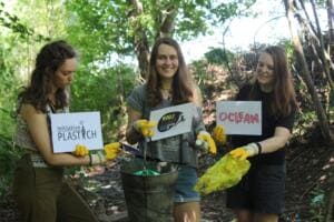 Drei junge Frauen mit Arbeitshandschuhen und Eimer halten Schilder mit den Aufschriften Initiative Plastich, World Cleanup Day und OClean in einem Waldstück.