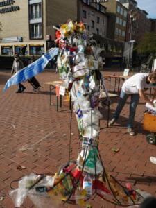 Eine hohe Skulptur aus gesammeltem Plastikmüll steht auf einem Backsteinplatz, im Hintergrund Häuser und ein Helfer.