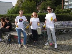 Drei junge Erwachsene im World-Cleanup-Day-Shirt zeigen vor einer Graffitimauer den Daumen hoch und halten Eimer und Greifzangen.
