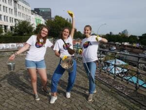 Drei junge Frauen im World-Cleanup-Day-Shirt jubeln mit Eimern voller Zigarettenstummel auf einem Platz über einer Straße.