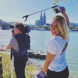 Eine lachende Frau im World-Cleanup-Day-Shirt hält eine Greifzange hoch, neben ihr filmt ein Kameramann, im Hintergrund Rhein und Kölner Dom.