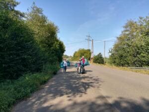 Zwei Personen tragen volle blaue Säcke und einen alten Reifen eine sonnige Landstraße zwischen Büschen entlang.