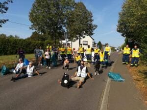 Eine große Gruppe in gelben Warnwesten sitzt und steht mit Säcken auf einer Straße am Ortsrand, vorne liegen Fundstücke.