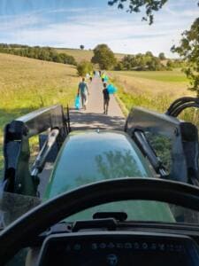 Blick aus der Kabine eines Traktors auf einen Feldweg, auf dem Helferinnen und Helfer mit blauen Säcken vorausgehen.