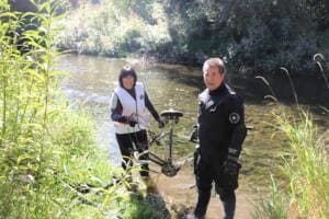 Eine Frau in World-Cleanup-Day-Weste und ein Mann im Trockenanzug stehen im Flussbett der Gera und halten ein gefundenes Fahrrad.