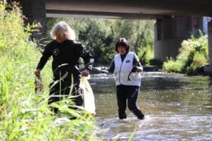 Zwei Frauen, eine im Trockenanzug, eine in World-Cleanup-Day-Weste, sammeln unter einer Brücke im Flussbett der Gera Müll.