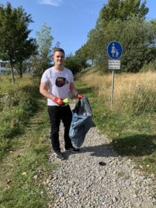 Ein junger Mann im World-Cleanup-Day-Shirt steht mit Müllsack und Greifzange auf einem Schotterweg in Oberhof.