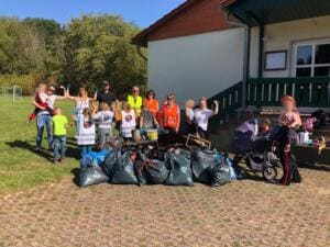 Eine Gruppe aus Erwachsenen und Kindern steht vor einem Gebäude hinter vielen vollen grauen Müllsäcken auf einem Platz.