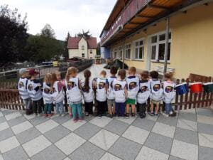 Eine Reihe Kindergartenkinder in World-Cleanup-Day-Westen steht von hinten vor einem Zaun auf einem Kita-Gelände.