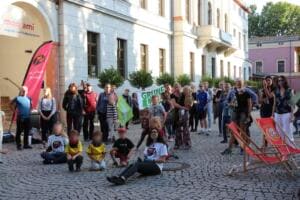 Eine große Gruppe aus Erwachsenen und Kindern sitzt und steht auf dem Platz vor dem Jugend- und Kulturzentrum mon ami in Weimar, daneben Liegestühle.