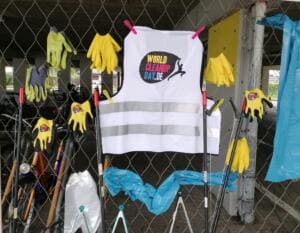 An einem Maschendrahtzaun hängen eine World-Cleanup-Day-Warnweste, gelbe Arbeitshandschuhe und Greifzangen.