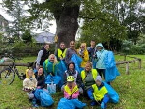 Eine Gruppe aus Erwachsenen und Kindern in Warnwesten sitzt und steht mit blauen Müllsäcken unter einem großen Baum auf einer Wiese.