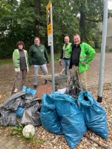 Vier Erwachsene, zwei davon in grünen Warnwesten, stehen an einem Parkplatzschild hinter vielen vollen blauen und grauen Müllsäcken und einer Schubkarre.