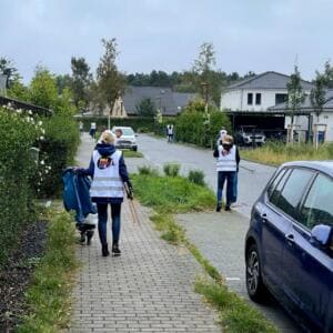 Zwei Frauen in World-Cleanup-Day-Warnwesten sammeln von hinten fotografiert mit Greifzangen und blauem Sack Müll entlang einer Wohnstraße mit Einfamilienhäusern.