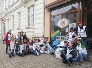 Eine Gruppe in World-Cleanup-Day-Westen sitzt und steht mit Greifzangen und vollen Säcken vor einem Ladengeschäft mit Regenbogenherz im Schaufenster.
