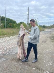 Ein junger Mann hält auf einem holprigen Weg neben Bahngleisen eine alte, verschmutzte Tischdecke hoch, die er an einem Stock gefunden hat.