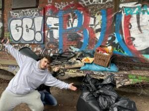 Ein junger Mann posiert mit ausgebreiteten Armen vor einer Graffitiwand, auf einem Haufen davor liegen ein Korb, Matratzenteile und volle Müllsäcke.