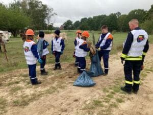 Kinder und Jugendliche der Jugendfeuerwehr in World-Cleanup-Day-Westen und Helmen sammeln mit Säcken Müll an einem Sandweg neben einer Kuhweide, ein Feuerwehrmann schaut zu.