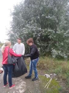 Eine Frau und zwei junge Männer halten an einem Wegrand einen großen schwarzen Müllsack auf, daneben liegen Holzlatten.