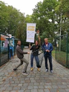 Drei Erwachsene posieren mit Greifzangen und einem Fahnenbanner mit der Aufschrift Litter Picker vor dem Eingang des Strandbads Plötzensee.