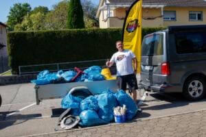 Ein Mann im World-Cleanup-Day-T-Shirt zeigt den Daumen hoch neben einem Anhänger voller blauer Müllsäcke, daneben eine gelbe World-Cleanup-Day-Beachflag.