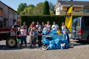 Eine Gruppe aus Kindern und Erwachsenen steht vor einem Traktor und einem Anhänger voller blauer Müllsäcke, daneben eine gelbe World-Cleanup-Day-Beachflag.