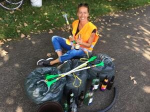Eine lachende Frau in orangefarbener Warnweste sitzt auf einem Weg zwischen vollen Müllsäcken, Glasflaschen und Greifzangen und hält eine Flasche hoch.