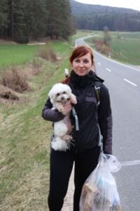 Eine Frau mit einem kleinen weißen Hund auf dem Arm und einem vollen Müllbeutel steht am Rand einer Landstraße.