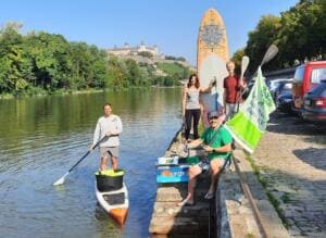 Ein Mann auf einem Stand-up-Paddle-Board mit Eimer paddelt auf dem Main, am Ufer stehen weitere Personen mit Boards und einer Fahne, im Hintergrund die Festung Marienberg.
