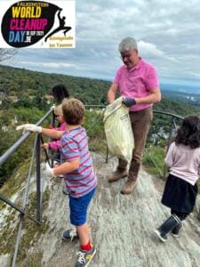 Ein Mann und drei Kinder sammeln auf einem Felsen mit Geländer Müll in einen Sack, im Hintergrund weite Landschaft; oben links ist ein World-Cleanup-Day-Logo eingeblendet.