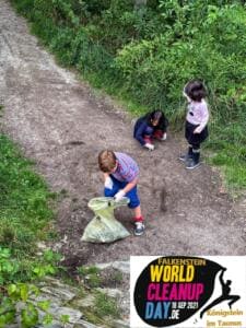 Drei Kinder sammeln von oben fotografiert auf einem Waldweg Müll in einen Beutel; unten rechts ist ein World-Cleanup-Day-Logo eingeblendet.