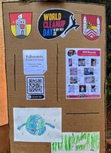 Eine Pappwand mit dem World-Cleanup-Day-Logo, zwei Ortswappen, Plakaten zum Müll-Museum, einem QR-Code und Kinderzeichnungen einer lachenden Erde.
