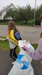 Ein Mädchen mit World-Cleanup-Day-Schärpe hält gemeinsam mit einer weiteren Person einen weißen Müllsack am Straßenrand auf.
