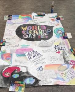Ein großes selbst gemaltes Plakat mit World-Cleanup-Day-Logo und Sprüchen wie Plastik zerstört die Meere liegt mit Farbkästen auf dem Boden.