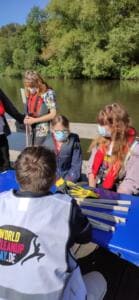 Schülerinnen und Schüler in Schwimmwesten sitzen mit Greifzangen in einem Boot auf einem Fluss, im Vordergrund ein Junge in World-Cleanup-Day-Weste.