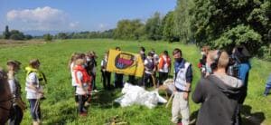 Schülerinnen und Schüler in Warnwesten und Schwimmwesten stehen mit einer gelben World-Cleanup-Day-Fahne auf einer Wiese, ein Kamerateam filmt.