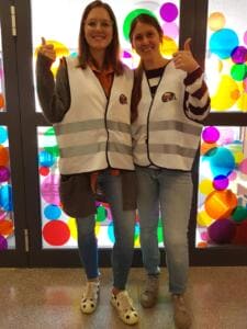 Zwei lächelnde Frauen in World-Cleanup-Day-Warnwesten zeigen vor einer bunt beklebten Glastür den Daumen hoch.