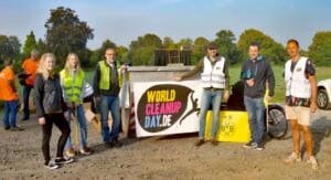 Neun Erwachsene in Warnwesten stehen bei Sonnenaufgang vor einem Container mit World-Cleanup-Day-Banner und BVB-Logo.