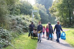 Eine Gruppe aus Erwachsenen und Kindern geht mit einem Handwagen, Greifzangen und blauen Säcken einen Radweg zwischen Büschen entlang.