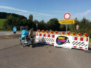 Ein World-Cleanup-Day-Banner hängt an einer rot-weißen Straßenabsperrung mit Warnleuchten, daneben ein Bollerwagen mit Säcken und ein Umleitungsschild.