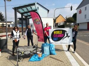 Vier Erwachsene stehen an einer Bushaltestelle mit World-Cleanup-Day-Banner, Beachflag, Bollerwagen und vollen blauen Säcken.