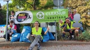 Eine Gruppe Erwachsener in Warnwesten sitzt und steht mit vollen blauen Säcken an einem Straßenrand vor einem grünen Banner mit der Aufschrift Cleanup.saarland.
