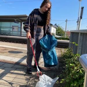 Eine junge Frau sammelt mit einer roten Greifzange und einem blauen Sack Müll an einem Bahnsteig.