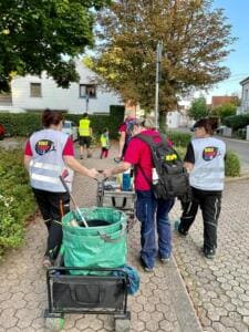 Drei Frauen in World-Cleanup-Day-Westen ziehen von hinten fotografiert einen Bollerwagen mit Gartensack einen Gehweg entlang.