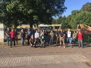 Eine große Gruppe aus Erwachsenen und Kindern steht im Sonnenschein auf einem Platz vor einem flachen Gebäude unter Bäumen.