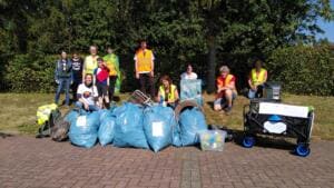 Eine Gruppe aus Erwachsenen und Kindern in Warnwesten steht und sitzt auf einer Wiese hinter vielen vollen blauen Säcken und einem Bollerwagen.